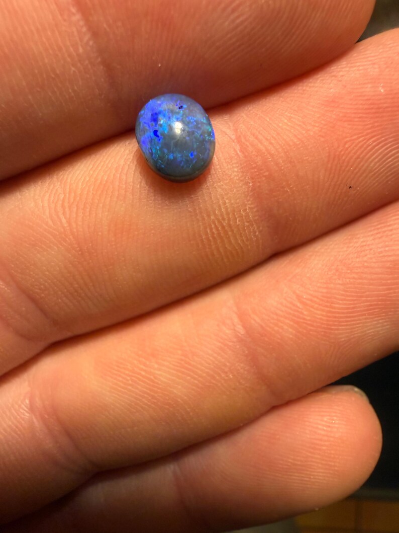 May include: A small, oval-shaped black opal gemstone featuring a dark base color with vibrant, iridescent flashes of bright blue and deep violet light reflecting across its polished, smooth, and rounded surface.