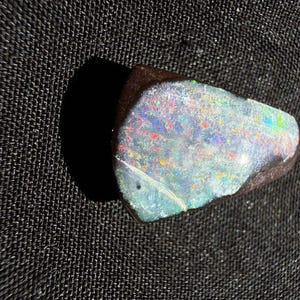 May include: A polished boulder opal gemstone with a dark brown ironstone matrix and a vibrant play-of-color surface featuring iridescent flashes of blue, green, and red light across its irregular, somewhat triangular shape.