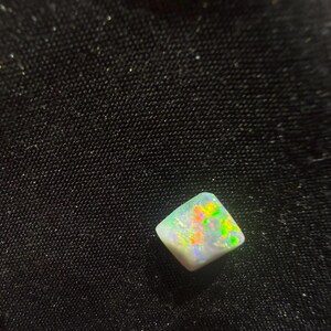 May include: A small, square-shaped opal gemstone featuring a white base color with vibrant, multi-colored flashes of green, yellow, and orange light reflecting across its polished, slightly irregular surface.