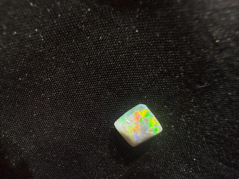 May include: A small, square-shaped opal gemstone featuring a white base color with vibrant, multi-colored flashes of green, yellow, and orange light reflecting across its polished, slightly irregular surface.
