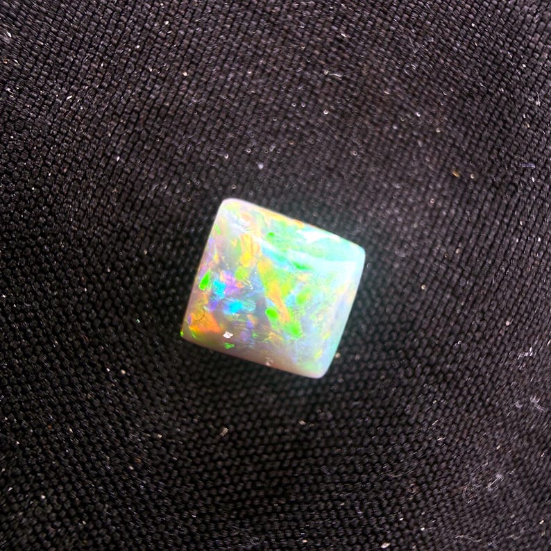 May include: A square-cut opal gemstone featuring a white base color with vibrant flashes of green, yellow, orange, and blue light. The stone has a smooth, polished surface that reflects light, showcasing a multi-colored play of color throughout.