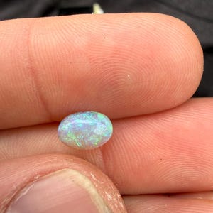 May include: An oval-shaped cabochon gemstone with a translucent white base and vibrant flashes of green and blue iridescence throughout the surface. The stone features a smooth, polished finish that reflects light, highlighting the internal play of color.