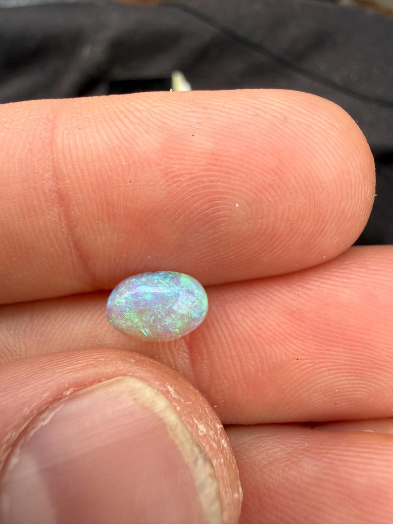 May include: An oval-shaped cabochon gemstone with a translucent white base and vibrant flashes of green and blue iridescence throughout the surface. The stone features a smooth, polished finish that reflects light, highlighting the internal play of color.