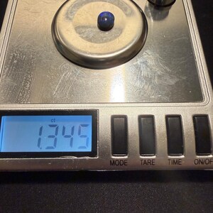 May include: A small, dark blue, oval-shaped gemstone sits on the circular weighing platform of a silver digital scale. The scale's blue backlit LCD screen displays the measurement of 1.345 ct, with a metal calibration weight visible nearby.