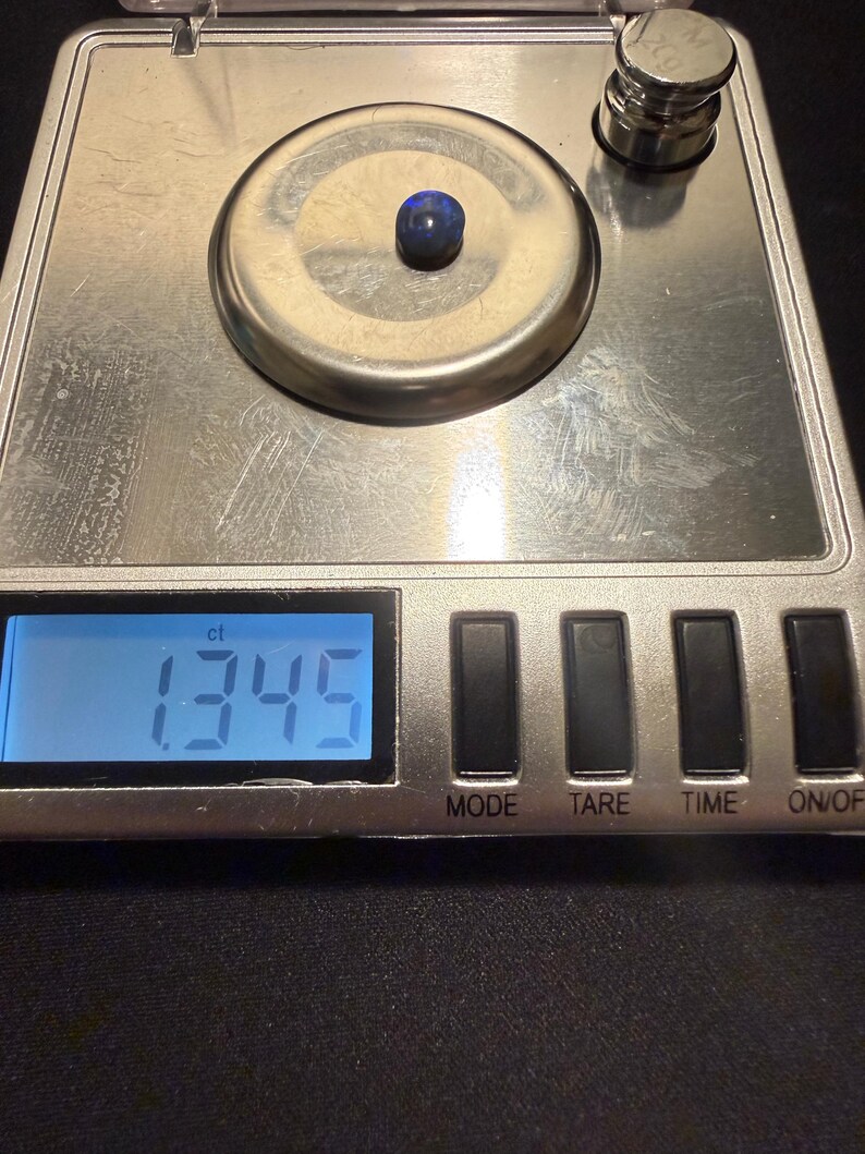 May include: A small, dark blue, oval-shaped gemstone sits on the circular weighing platform of a silver digital scale. The scale's blue backlit LCD screen displays the measurement of 1.345 ct, with a metal calibration weight visible nearby.