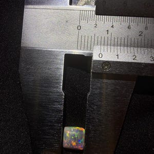 May include: A square-cut opal gemstone displaying iridescent flashes of red, green, and blue, positioned within the jaws of a metal caliper. The measurement tool indicates the stone has a width of approximately 0.35 inches.