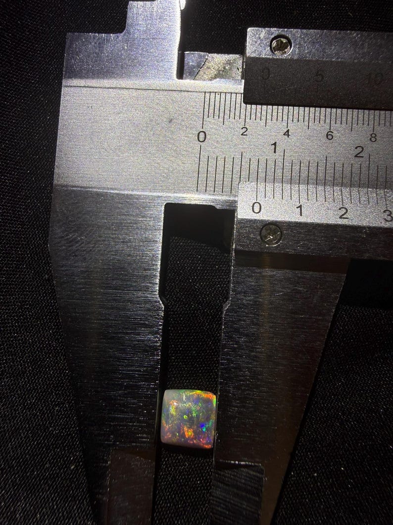 May include: A square-cut opal gemstone displaying iridescent flashes of red, green, and blue, positioned within the jaws of a metal caliper. The measurement tool indicates the stone has a width of approximately 0.35 inches.