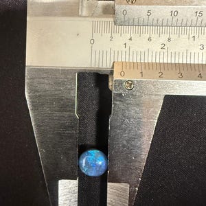 May include: A small, round, iridescent blue and black gemstone is positioned between the jaws of a metal caliper. The measuring tool indicates the stone has a diameter of approximately 0.35 inches, showing vibrant blue flashes against a dark base.