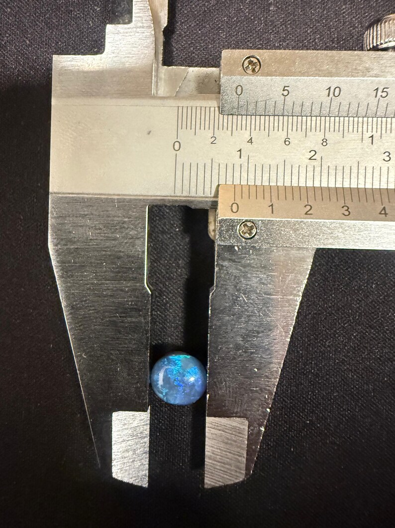 May include: A small, round, iridescent blue and black gemstone is positioned between the jaws of a metal caliper. The measuring tool indicates the stone has a diameter of approximately 0.35 inches, showing vibrant blue flashes against a dark base.