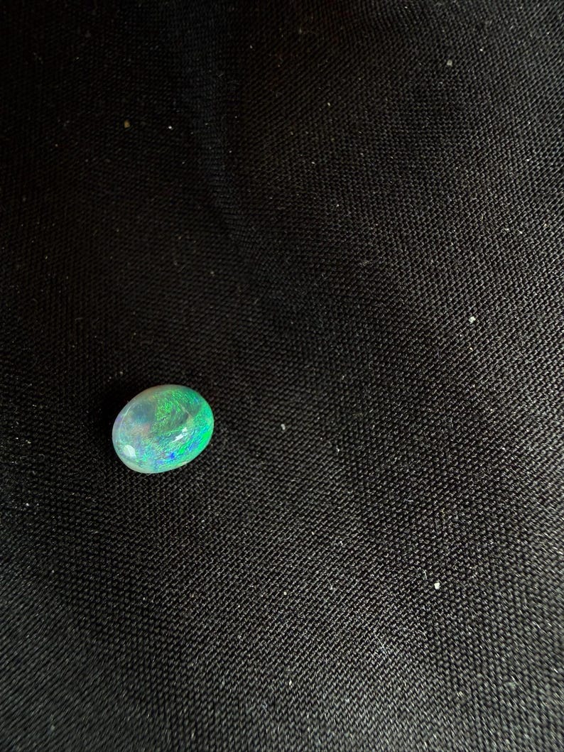 Peut inclure: Une pierre pr&eacute;cieuse opale de forme ovale lisse, pr&eacute;sentant une base translucide avec des &eacute;clats vibrants de lumi&egrave;re verte et bleue. La pierre affiche une surface polie et arrondie qui refl&egrave;te la lumi&egrave;re, r&eacute;v&eacute;lant un jeu de couleurs distinct.