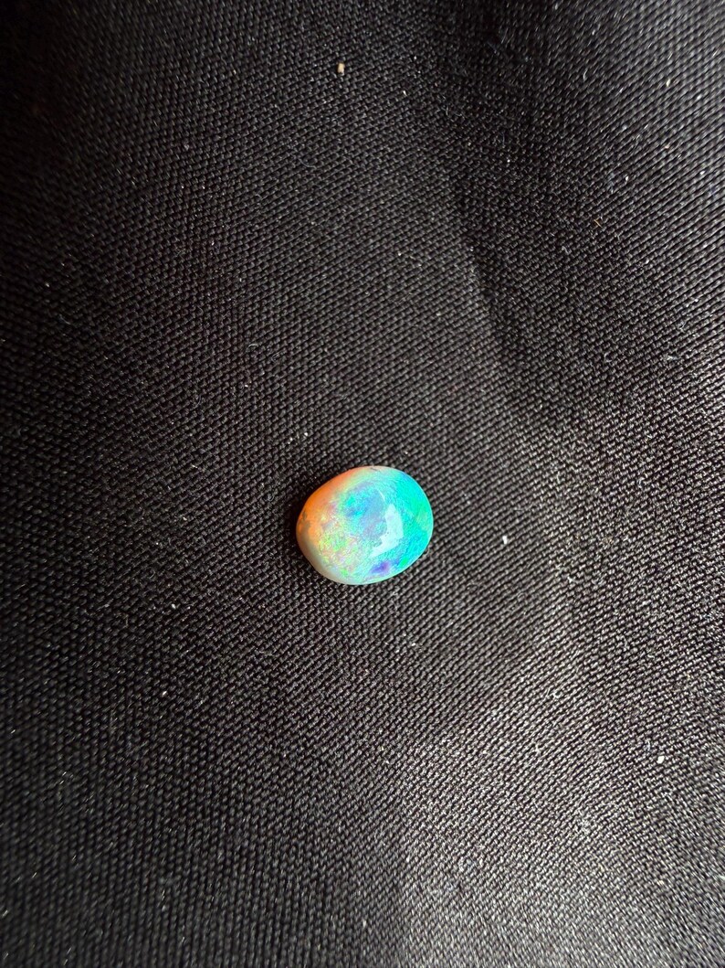 Peut inclure: Une pierre pr&eacute;cieuse opale de forme ovale et lisse pr&eacute;sentant un jeu de couleurs vibrant avec des &eacute;clats iridescents orange, verts et bleus sur sa surface polie. La pierre poss&egrave;de une qualit&eacute; translucide et une taille cabochon arrondie.