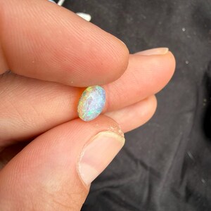 May include: A small, oval-shaped opal gemstone featuring a faceted surface that displays a vibrant play of color, including flashes of green, blue, and pink, held between two fingers against a dark background.
