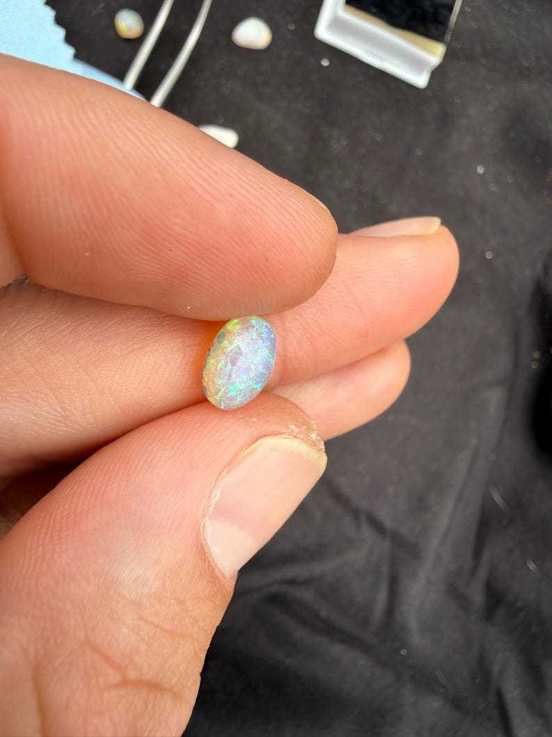 May include: A small, oval-shaped opal gemstone featuring a faceted surface that displays a vibrant play of color, including flashes of green, blue, and pink, held between two fingers against a dark background.