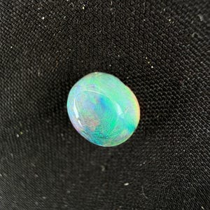 Peut inclure: Une pierre pr&eacute;cieuse opale lisse de forme ovale pr&eacute;sentant un jeu de couleurs vibrant avec des &eacute;clats iridescents de vert n&eacute;on, de bleu &eacute;lectrique et de l&eacute;g&egrave;res touches de rose et d'orange sur sa surface polie et translucide.