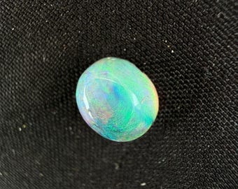 May include: A smooth, oval-shaped opal gemstone displaying a vibrant play-of-color with iridescent flashes of neon green, electric blue, and subtle hints of pink and orange across its polished, translucent surface against a dark textured background.