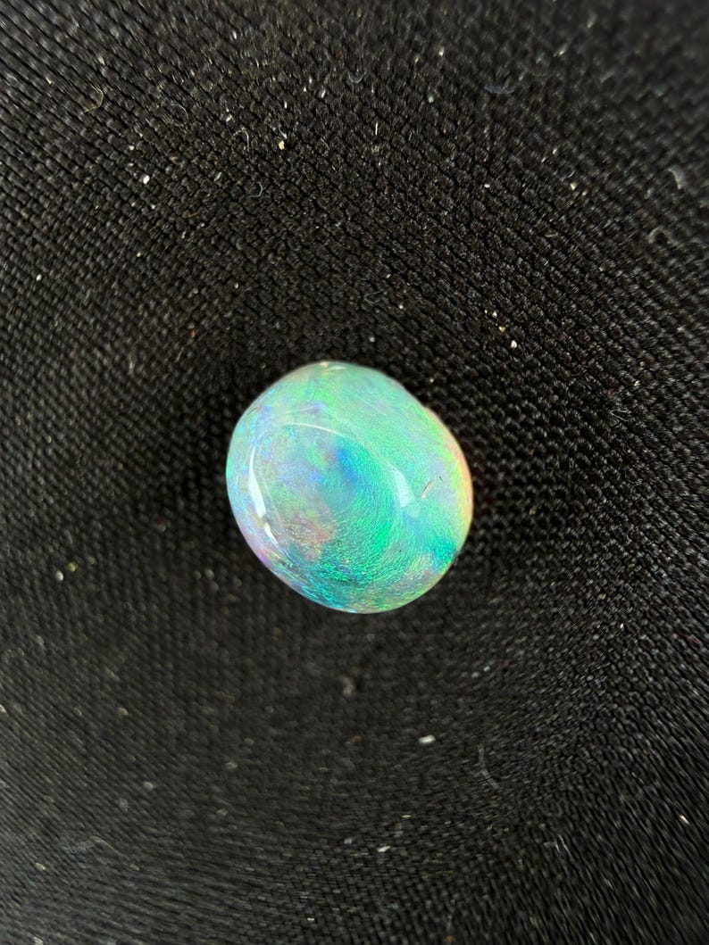 Peut inclure: Une pierre pr&eacute;cieuse opale lisse de forme ovale pr&eacute;sentant un jeu de couleurs vibrant avec des &eacute;clats iridescents de vert n&eacute;on, de bleu &eacute;lectrique et de l&eacute;g&egrave;res touches de rose et d'orange sur sa surface polie et translucide.