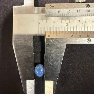 May include: An oval-shaped blue opal gemstone with iridescent flashes of color held within the jaws of a metal caliper. The measuring tool indicates the stone has a width of approximately 0.35 inches.