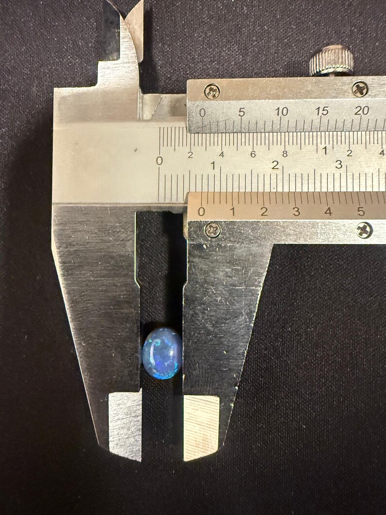 May include: An oval-shaped blue opal gemstone with iridescent flashes of color held within the jaws of a metal caliper. The measuring tool indicates the stone has a width of approximately 0.35 inches.
