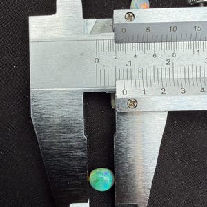 Peut inclure: Une opale ronde et iris&eacute;e aux reflets verts et bleus vifs, plac&eacute;e entre les m&acirc;choires d'un pied &agrave; coulisse en m&eacute;tal. Le pied &agrave; coulisse indique un diam&egrave;tre d'environ 8 millim&egrave;tres pour cette pierre pr&eacute;cieuse.