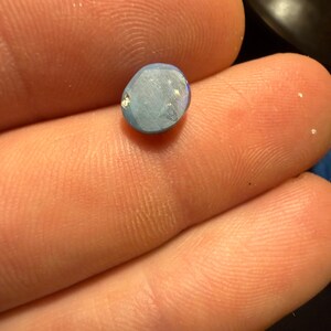 May include: A small, oval-shaped, light blue gemstone with a smooth, polished surface. The stone displays subtle iridescent flashes and a slightly irregular, rounded edge, reflecting light across its flat, matte-textured face.