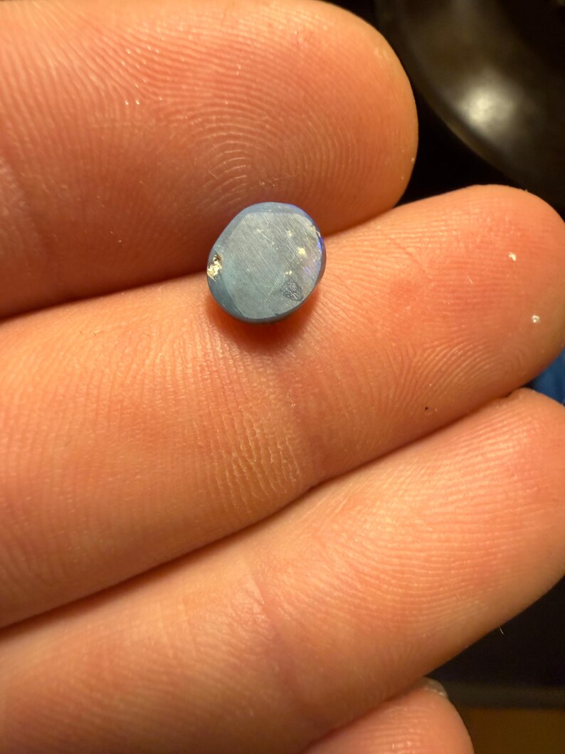 May include: A small, oval-shaped, light blue gemstone with a smooth, polished surface. The stone displays subtle iridescent flashes and a slightly irregular, rounded edge, reflecting light across its flat, matte-textured face.