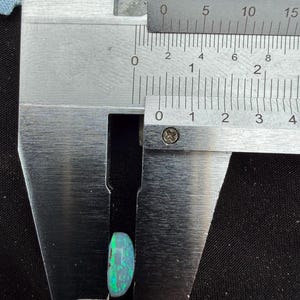 May include: A polished, oval-shaped opal gemstone with vibrant green and blue flashes of color. The gemstone is positioned within the jaws of a stainless steel caliper, which indicates a width of approximately 0.35 inches on the imperial scale.