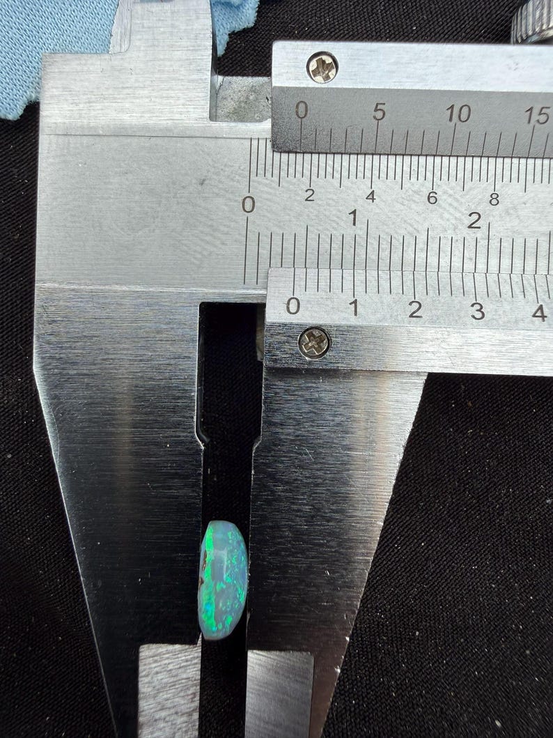 May include: A polished, oval-shaped opal gemstone with vibrant green and blue flashes of color. The gemstone is positioned within the jaws of a stainless steel caliper, which indicates a width of approximately 0.35 inches on the imperial scale.