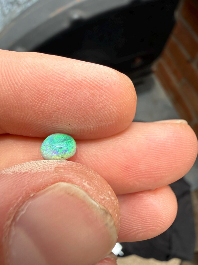 Peut inclure: Une petite pierre pr&eacute;cieuse polie et arrondie pr&eacute;sentant un jeu de couleurs vibrant avec des &eacute;clats iris&eacute;s de vert vif, de bleu &eacute;lectrique et de l&eacute;g&egrave;res nuances de violet sur sa surface lisse et bomb&eacute;e, tenue entre deux doigts.