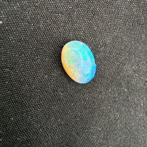 May include: An oval-shaped opal gemstone featuring a smooth, polished surface with a play-of-color effect. The stone displays vibrant flashes of blue, green, and orange hues across its translucent body, creating a multi-colored appearance.