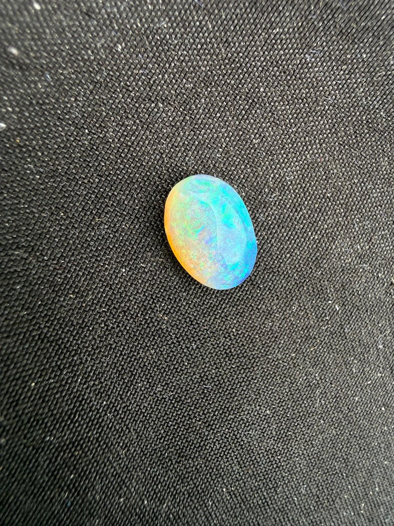 May include: An oval-shaped opal gemstone featuring a smooth, polished surface with a play-of-color effect. The stone displays vibrant flashes of blue, green, and orange hues across its translucent body, creating a multi-colored appearance.