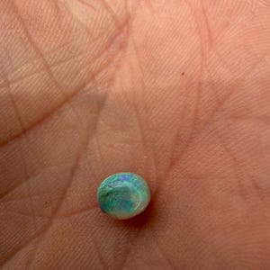 Peut inclure: Une petite pierre pr&eacute;cieuse opale polie, de forme ronde, pr&eacute;sentant une couleur de base blanche avec des &eacute;clats iris&eacute;s vibrants de vert et de bleu sur sa surface. La pierre a une forme cabochon lisse et bomb&eacute;e qui refl&egrave;te la lumi&egrave;re.