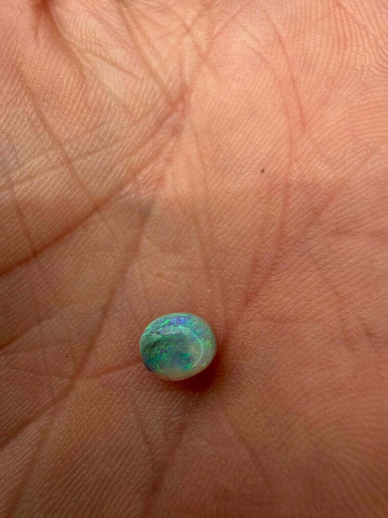 Peut inclure: Une petite pierre pr&eacute;cieuse opale polie, de forme ronde, pr&eacute;sentant une couleur de base blanche avec des &eacute;clats iris&eacute;s vibrants de vert et de bleu sur sa surface. La pierre a une forme cabochon lisse et bomb&eacute;e qui refl&egrave;te la lumi&egrave;re.