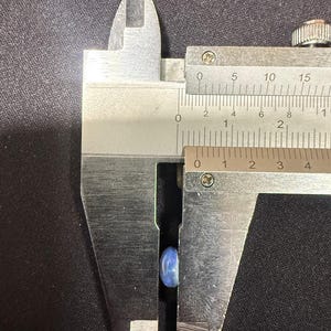 May include: A small, smooth, light blue oval-shaped stone is positioned between the jaws of a metal caliper. The measuring tool indicates the stone has a width of approximately 0.3 inches.