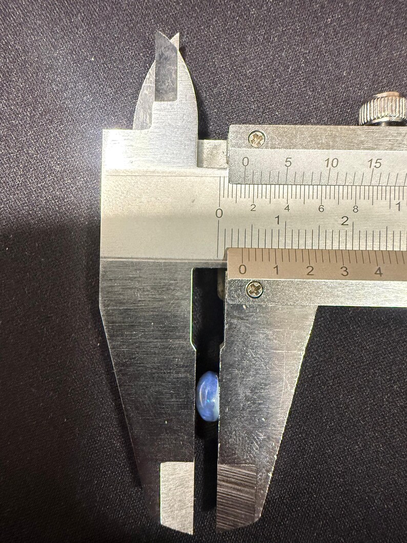 May include: A small, smooth, light blue oval-shaped stone is positioned between the jaws of a metal caliper. The measuring tool indicates the stone has a width of approximately 0.3 inches.