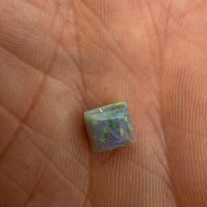 May include: A square-cut opal gemstone featuring a dark grey base color with vibrant flashes of green, blue, and purple light reflecting across its polished surface, displaying a distinct play-of-color pattern throughout the stone.