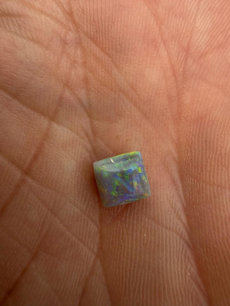 May include: A square-cut opal gemstone featuring a dark grey base color with vibrant flashes of green, blue, and purple light reflecting across its polished surface, displaying a distinct play-of-color pattern throughout the stone.
