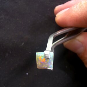 May include: A square-cut opal gemstone with a white base and vibrant flashes of orange, green, and blue color play. The gemstone is held by a pair of silver-toned metal tweezers, showcasing its polished surface and distinct iridescent internal patterns.