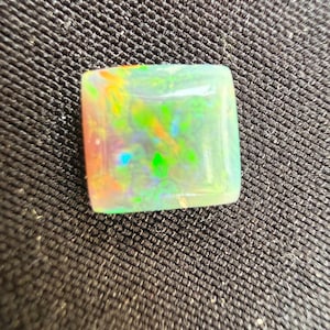 May include: A square-cut opal gemstone featuring a translucent white base with vibrant flashes of neon green, orange, and blue play-of-color throughout the surface. The stone has a smooth, polished finish and a flat top with slightly rounded edges.
