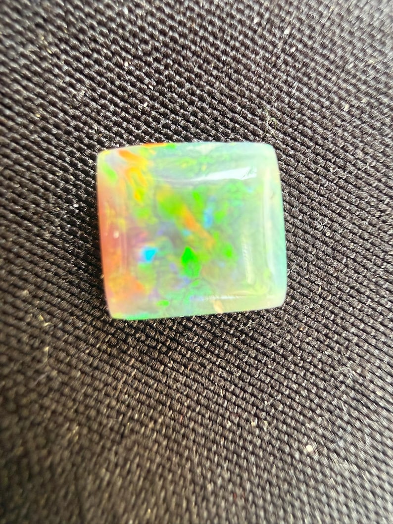 May include: A square-cut opal gemstone featuring a translucent white base with vibrant flashes of neon green, orange, and blue play-of-color throughout the surface. The stone has a smooth, polished finish and a flat top with slightly rounded edges.