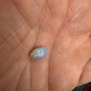 May include: An oval-shaped opal gemstone displaying a play-of-color with flashes of blue, green, and white light. The stone has a smooth, polished surface and a translucent appearance, showcasing internal color variations across its rounded form.