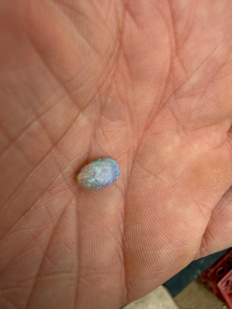 May include: An oval-shaped opal gemstone displaying a play-of-color with flashes of blue, green, and white light. The stone has a smooth, polished surface and a translucent appearance, showcasing internal color variations across its rounded form.