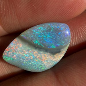 May include: A polished, teardrop-shaped opal gemstone featuring a vibrant play-of-color with flashes of bright green, electric blue, and fiery orange against a light, semi-translucent base. The stone has a smooth, undulating surface texture.