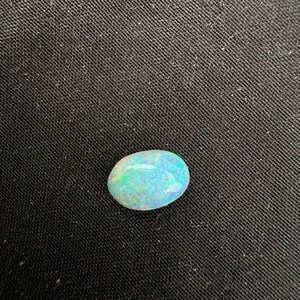 May include: An oval-shaped cabochon gemstone with a smooth, polished surface. The stone displays a translucent white base color with vibrant flashes of iridescent green and blue light reflecting across its rounded, dome-like top surface.