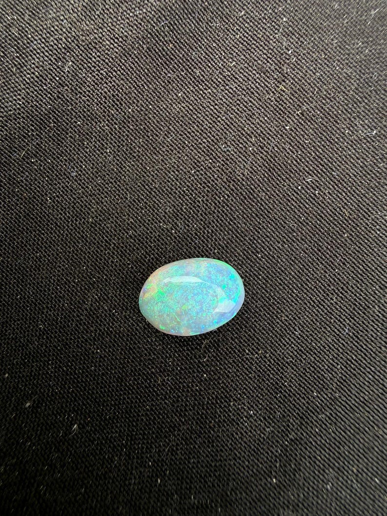 May include: An oval-shaped cabochon gemstone with a smooth, polished surface. The stone displays a translucent white base color with vibrant flashes of iridescent green and blue light reflecting across its rounded, dome-like top surface.