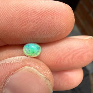 Peut inclure: Une petite pierre pr&eacute;cieuse en cabochon de forme ovale avec une base blanche translucide et des &eacute;clats vibrants de lumi&egrave;re verte et bleue iris&eacute;e. La pierre pr&eacute;sente un effet de jeu de couleurs sur sa surface lisse et polie, refl&eacute;tant la lumi&egrave;re dans diverses teintes vives et satur&eacute;es.