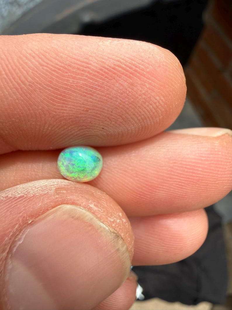 Peut inclure: Une petite pierre pr&eacute;cieuse en cabochon de forme ovale avec une base blanche translucide et des &eacute;clats vibrants de lumi&egrave;re verte et bleue iris&eacute;e. La pierre pr&eacute;sente un effet de jeu de couleurs sur sa surface lisse et polie, refl&eacute;tant la lumi&egrave;re dans diverses teintes vives et satur&eacute;es.