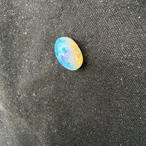 May include: An oval-shaped opal gemstone displaying a play-of-color with iridescent flashes of blue, green, and orange against a translucent, milky white base. The stone has a smooth, polished cabochon surface that reflects light.