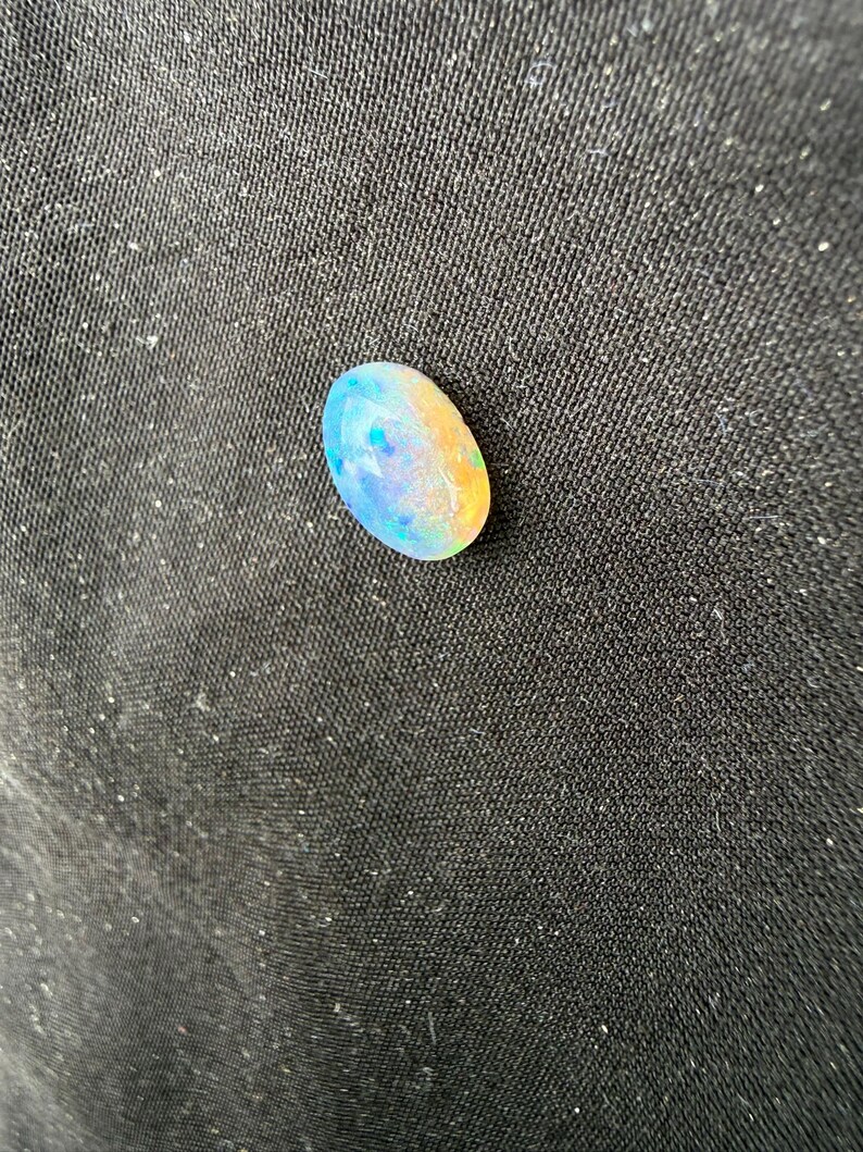 May include: An oval-shaped opal gemstone displaying a play-of-color with iridescent flashes of blue, green, and orange against a translucent, milky white base. The stone has a smooth, polished cabochon surface that reflects light.