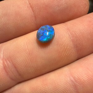 May include: A small, oval-shaped black opal gemstone featuring a polished, smooth surface with vibrant, iridescent flashes of electric blue and deep green light reflecting from within the dark, opaque stone structure.