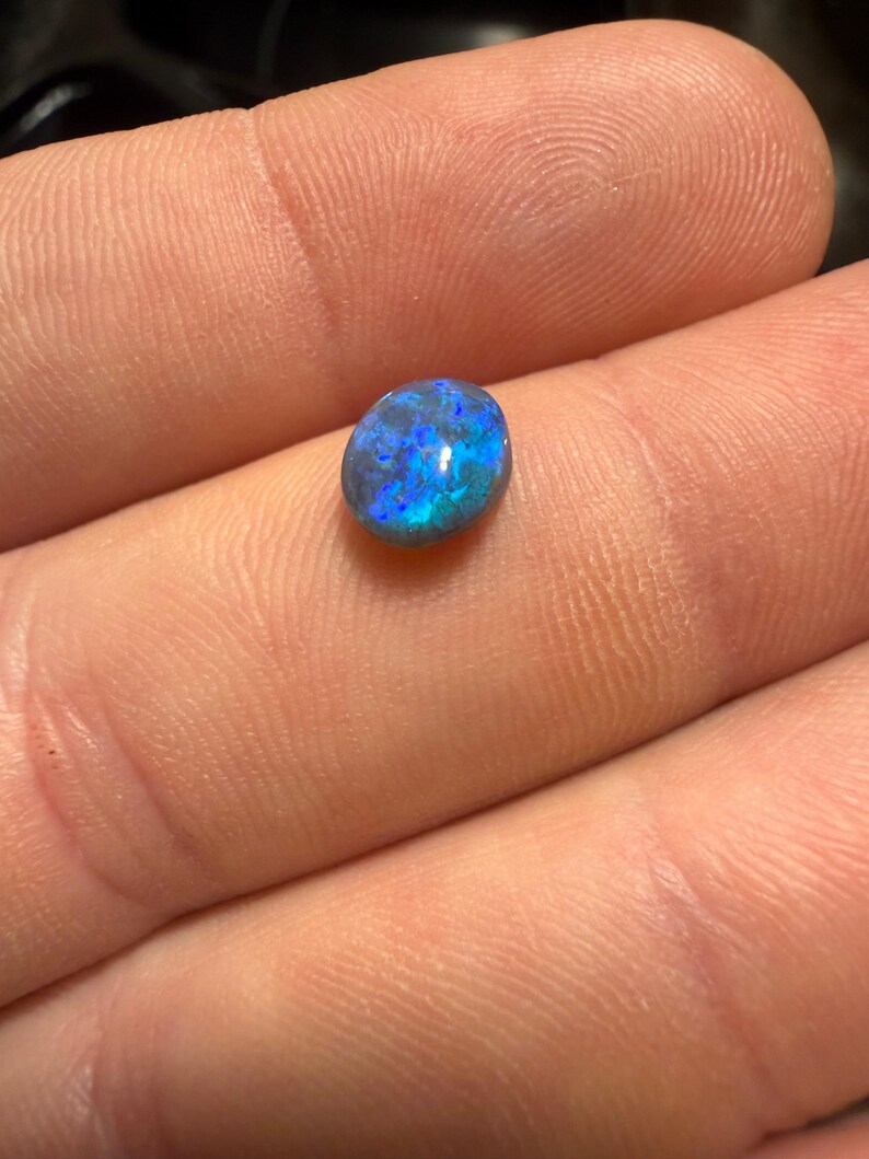 May include: A small, oval-shaped black opal gemstone featuring a polished, smooth surface with vibrant, iridescent flashes of electric blue and deep green light reflecting from within the dark, opaque stone structure.